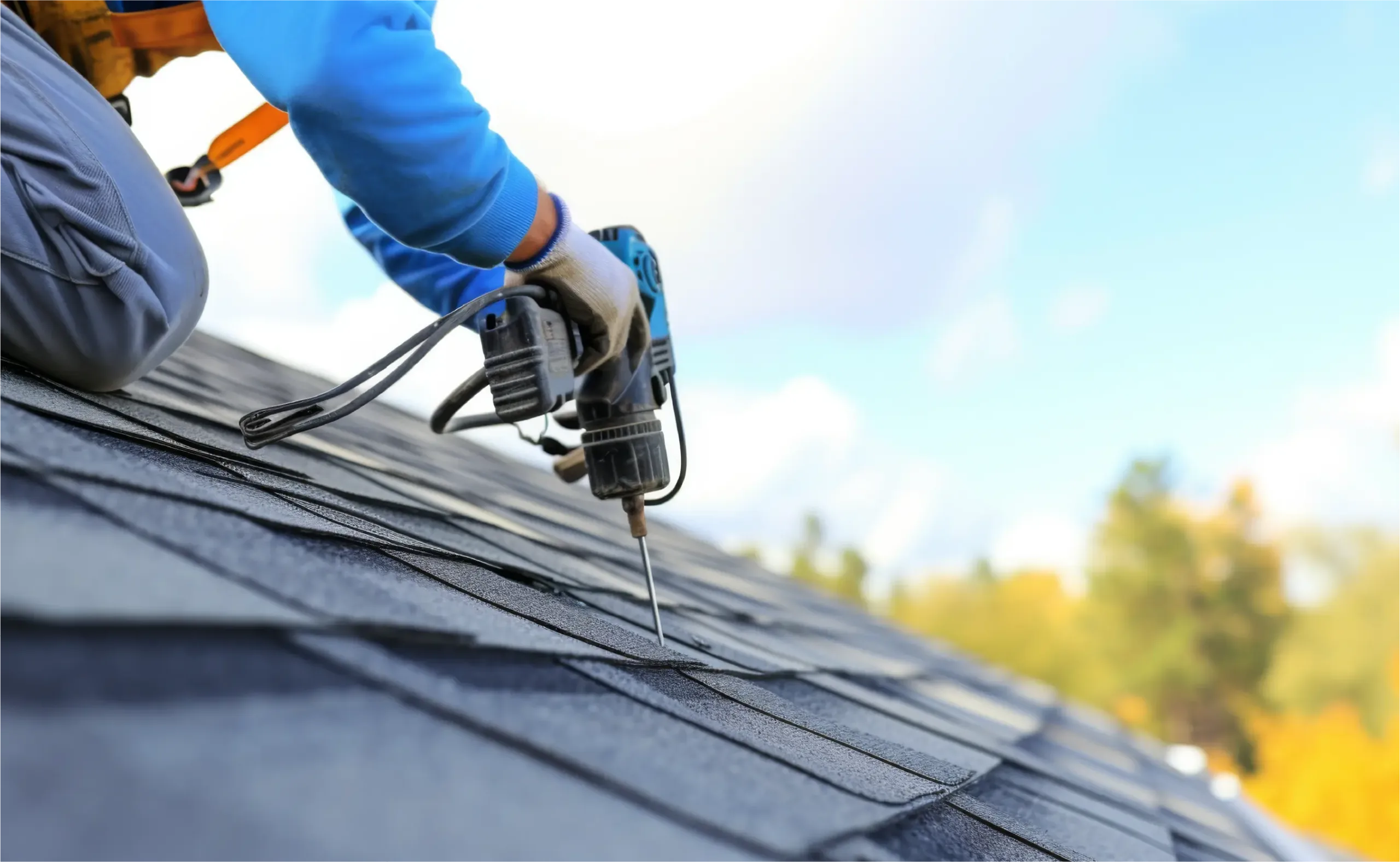 A roofer wearing safety gear uses a power drill to install asphalt shingles on a residential roof under a blue sky.